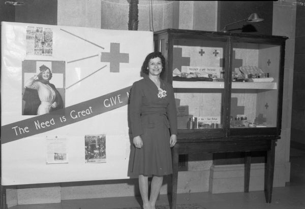Ruth Nelson, Red Cross motor corps volunteer, and the Red Cross display in the Wisconsin State Capitol rotunda. Display features contents of the prisoner-of-war capture parcel and weekly food package supplied to captured service men by the International Red Cross.