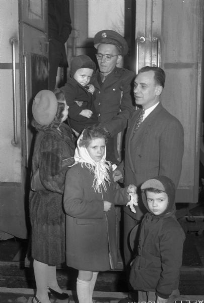 Corporal William (Bill) Lovelace, 319 North Blair Street, returning home after spending one year hiding from the Japanese and two years in a Japanese prisoner-of-war camp in Manila, Phillippines. He is being greeted at the railway station in Madison by his parents and siblings.  Top, left to right are: Mrs. W.I. (Gladys) Lovelace, Corporal Lovelace holding his brother Gary whom he has never seen, and Mr. Wilber I. Lovelace. Bottom row, left to right are: sister Joyce and brother Bobby.