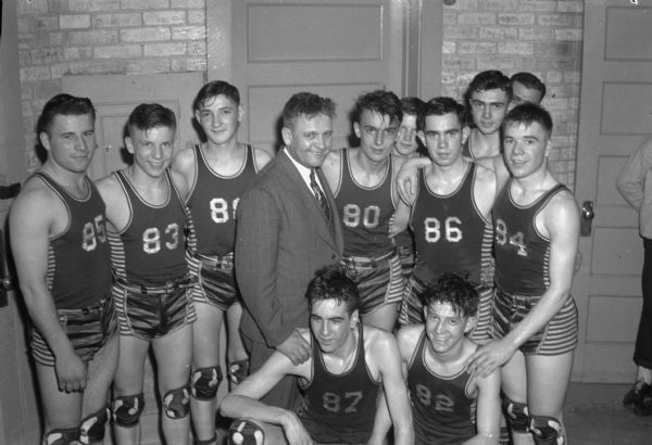 Group portrait of Lena High School's basketball team (Wildcats) taken at the annual state basketball tournament in Madison.  Kneeling left to right are:  Clarence Blahnik and Roman Kosner.  Standing left to right are:  Jack Kowalczyk, Marvin Christropherson, Lenorad Belongia, coach Bruce L. Kimball, Jerome Pelishek, Maynard Ausloos, and Arvid McGuire.  In the right background barely visible are: Francis De Jariais and L. Skarda.
