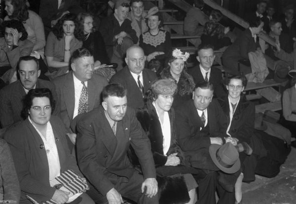 Parent of four of the players of the Lena High School basketball team seated in the stands during the annual state basketball tournament.  Front row left to right:  Mrs. John Ausloos, Gus Blahnik, John McGuire, and Mrs. McGuire.  Back row left to right are:  John Ausloos, Mrs. Emil Kosner and Mr. Kosner.