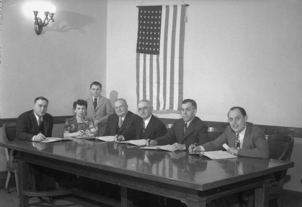 Group portrait of the Wisconsin Senate Veterans' & Military Affairs Committee sitting around a table with a Unites States flag in the background.