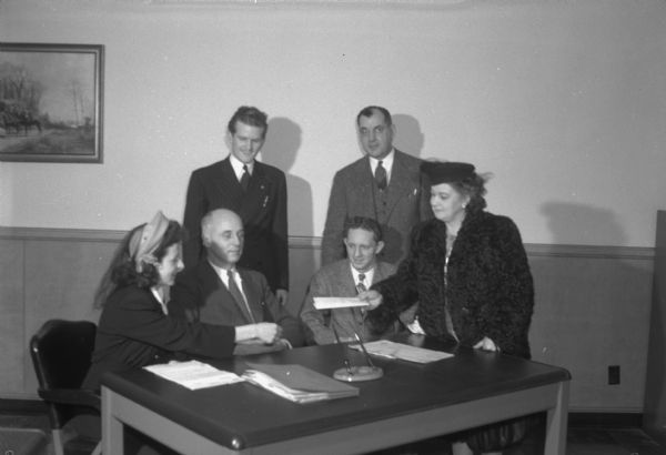 Anchor Savings and Loan Association staff making the first G.I. loan to Arthur L. Cass, discharged veteran, and his wife so they can purchase a home at 3305 Oak Court, in the Village of Monona. Left to right: Mrs. Cass, J.H. Ottow, assistant secretary; Mr. Cass; standing left to right: Guy S. Williams, local contact representative of Madison Veterans Office; A.C. Steinhauer, secretary-treasurer of Anchor; Mrs. George Post, seller of property, shown handing the deed to Mrs. Cass.