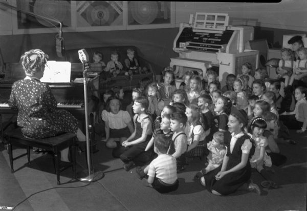 Group of children seated on the floor at the WHA radio studio listening to a woman (Fanny Steve?) play the piano during a program called "Fun Time".
