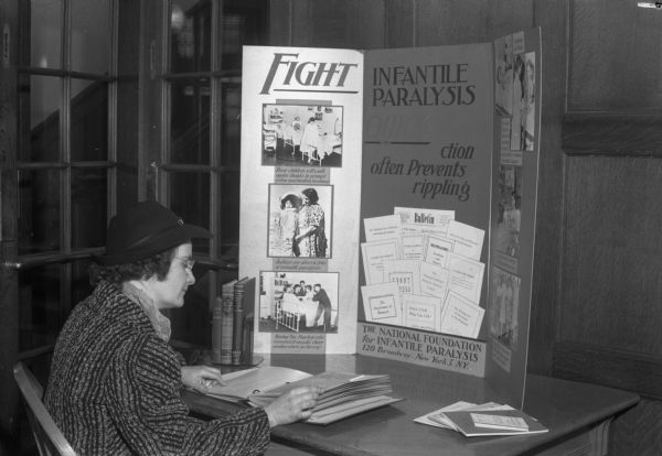 A women seated at a desk with a display board titled "Fight Infantile Paralysis".