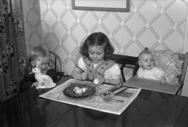 Janet Oldenburg decorating Easter eggs as her sister Nancy is watching. An infant is sitting in a high chair on the right. They are the children of Mr. and Mrs. Hugh (Elizabeth) Oldenburg, 3429 Crestwood Drive, Shorewood Hills.