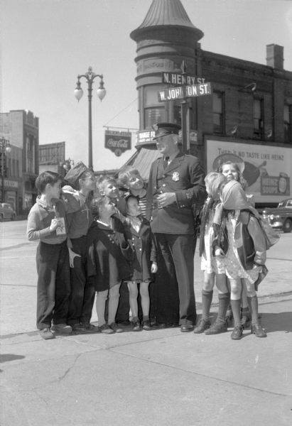 Officer Frank B. Ingraham, at his beat on the corners of State, Henry, and Johnson Streets, with young school children from Holy Redeemer School. Ingraham is retiring after 22 years with the Madison Police Department. The girl with pig-tails standing second to the left of Officer Ingraham is Sharon Ann Lewis, daughter of PFC. Narvil and Leona Lewis.