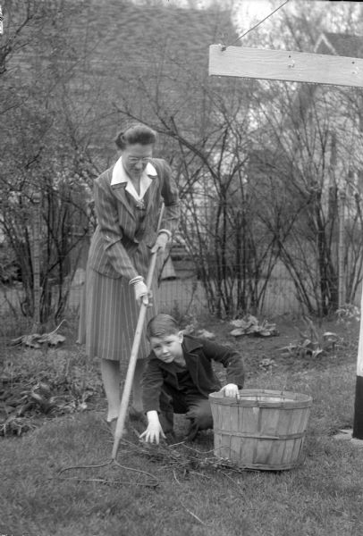 Mrs. Lillian Helfrecht raking her lawn with the help of her neighbor, six-year-old George Armstrong. George helped Mrs. Helfrecht with various chores because her three sons were away in military service. One of her sons, 1st Lieut. Kenneth Helfrecht, had an image of George painted on his Mustang fighter plane in appreciation for George's help.