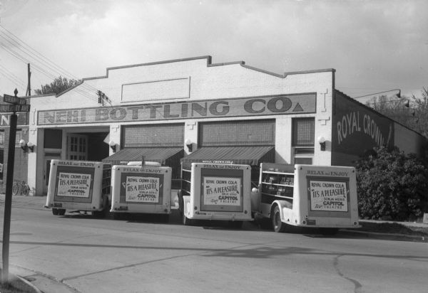 Company trucks in front of Nehi Bottling Company (Royal Crown Soda), 2600 University Avenue at the intersection of Grand Avenue. The back ends of the four trucks advertise the movie "It's A Pleasure" playing at the Capitol Theatre.