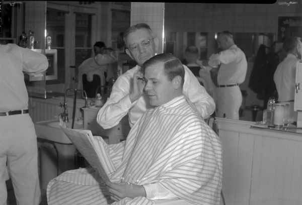 Barber giving haircut to a man reading a newspaper announcing Allied Forces victory in Europe.