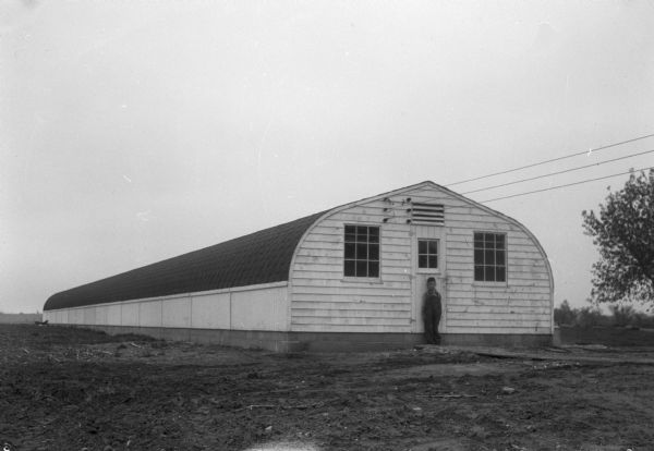 View across field towards a boy standing at the door to a brooder house. The building is 18 feet x 180 feet, complete with hot water heating, on the Frank Lyons turkey farm on Main Street, Verona.