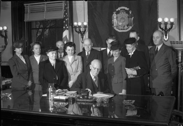 Governor Goodland signing a bill (probably, Ch 131, Laws of 1945 relaxing the state's food sanitation law and easing animal slaughter on family farms) with eleven onlookers.