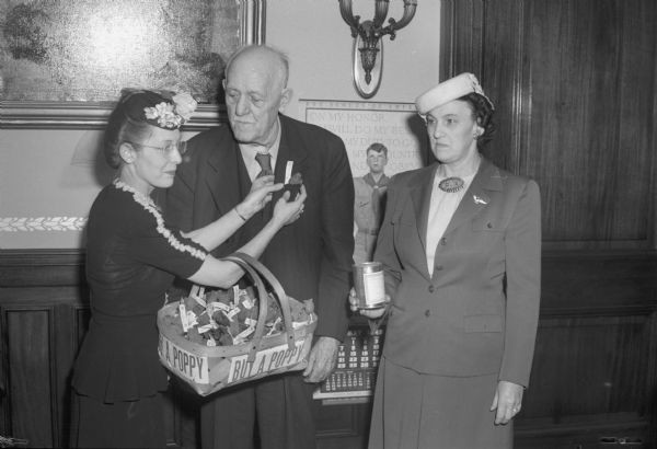 Governor Walter S. Goodland buying the first poppy from American Legion Auxiliary and Veterans of Foreign War Auxiliary members. Pictured left to right: Mrs. Mary Ellen Hawley, America Legion Auxiliary, pinning a poppy on Governor Goodland, center, and Mrs. Frieda Strassburg, right, Veterans of Foreign Wars Auxiliary, looking on.
