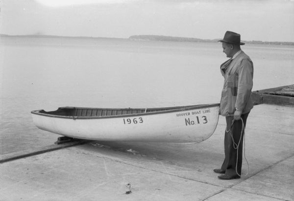 Bennie Berg standing on a lake shore dock next to a Hoover Boat Line boat. Also shows his invention to aid in launching, the Berg's Inc. Boat Roller.
