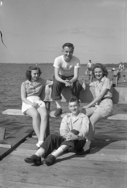 Group portrait of four Madison Central High School students relaxing on a pier on Lake Mendota. Pictured left to right: Alice Jensen, daughter of Mr. and Mrs. Otto H. Jensen, 413 Washburn Place; Dick Price, son of Mrs. Charlotte Price, 651 University Avenue; Jannette Lindauer, daughter of Mr. and Mrs. A.C. Lindauer, 310 North Hamilton Street; and Eugene Osborn, (seated on the pier), son of Mr. and Mrs. Eugene E. Osborn, 245 Langdon Street.