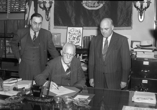 Wisconsin Governor Walter S. Goodland signing a bill in his office, with two men looking on. The bill is probably Chapter 233 of the Laws of 1945 on the assembling and publication of agricultural statistics.