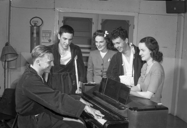Group portrait of Red Cross sponsored group, "Parlor Tricks," who entertain convalescent patients in the Army Air Force's regional hospital at Truax Field. Seated at the piano is Sgt. Howard Olson, entertaining some of his fellow GI fellow patients and "Parlor Tricks" members. Standing left to right: Pvt. David Miller, Chicago, Illinois; Betty Isabella, 2245 Eton Ridge; Pvt. Thomas Faile, White Plains, New York; and Mary Frances Dolan, 437 West Mifflin Street.