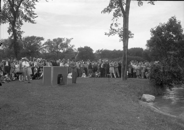 Onlookers near a pond at the Retriever Dog Field Trials in Vilas Park. James Simpson, Jr., of Chicago, one of the judges, has his hand upraised to signal; Leroy Beardsley of Chicago, another judge, is at the left with his arms folded. In the blind with his Irish Water Spaniel, "Big Mike Mahoney", is William Raymond of Fond du Lac. From the newspaper caption: Two Madison dogs, Stark Lester's "Buck of Monona" and Tony Bonovich's "Nigger of Upham Street" took top prizes.