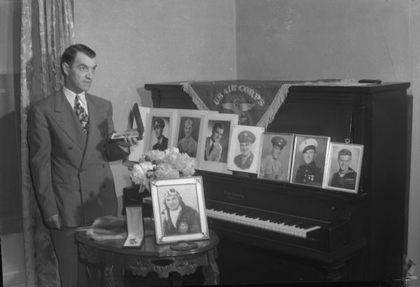 Fred M. Hoffman standing by a piano with photographs of seven of his sons, and a U.S. Air Corps banner. Five are still in the military service, two received medical discharges and one, Capt. Sylvester "Bud" Hoffman, was killed. On a table in front of the piano are flowers, a photograph of Bud, and the air medal and purple heart Bud was awarded. A ninth son, Thomas, was too young to serve in World War II.