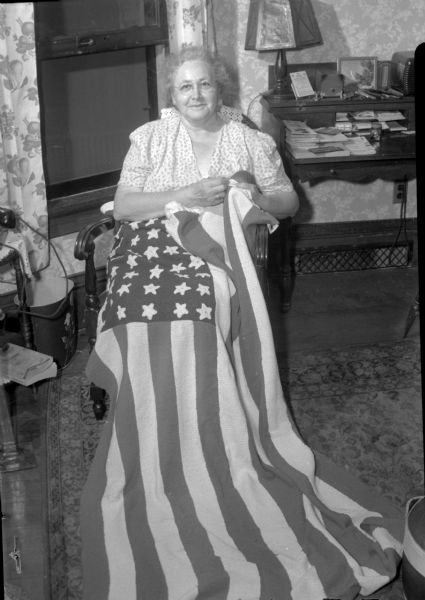 Mrs. Charles C. (Alice) Long is shown with the flag she crocheted for her son, Lieutenant Commander H.W. Christensen, who is a surgeon, serving in the Pacific area. The flag measures three feet ten inches by six feet, and took six months for her to make. It will be hung in the doctor's office when he returns from the war.