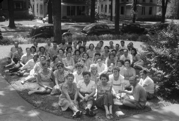 Group portrait of students seated on the front lawn next to the street. The Abraham Lincoln School for Social Science, Chicago, Illinois, sponsored a Summer Institute in Madison for several summers, including 1945. The students who attended the two week sessions were mostly adult union workers and their families. They lived in rooming houses on Langdon Street and Lake Lawn Place, attended lectures and participated in recreational activities. The photographs show the teachers with groups of students, both indoors and out-of-doors, the students participating in recreational activities such as boating and badminton and the children with a day care worker. The School was run by former University of Wisconsin Associate Professor of Classics Alban D. Winspear. Their textbook was called "Why Work for Nothing" espousing Marx's theory of value.  The school had a communist / leftist philosophy and was often confused with the University of Wisconsin's School for Workers.