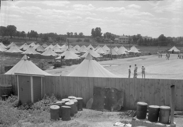 German prisoner of war camp with tents and fencing surrounding the area. Several people are on the grounds. Prisoners are working in the local cannery.