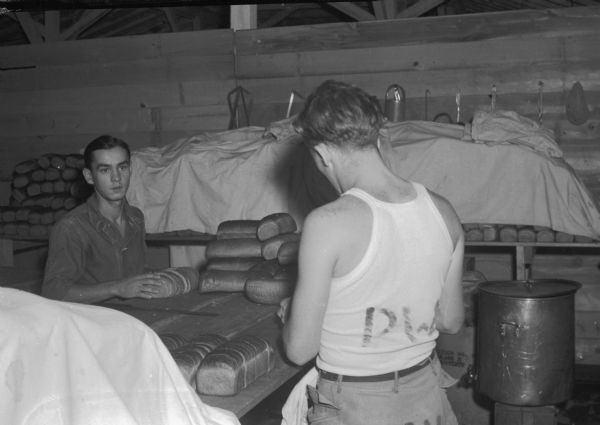 Two German prisoners of war, at a prisoner of war camp, cutting rye bread. The food preparation area includes large quantities of bread on another table covered with a tarpaulin. The camp had a population of 500 prisoners.