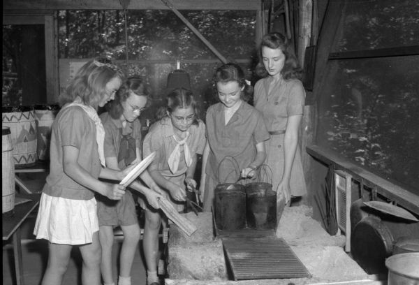 Four girl scouts and a camp counselor starting a fire in a camp fireplace as they start to prepare dinner at Camp Brandenburg near Springfield Corners.  Pictured from left to right:  Priscilla Nelson, Marie Anthony, Jane Hampton, Nancy Hughes, and camp councelor Betty Kletzien.