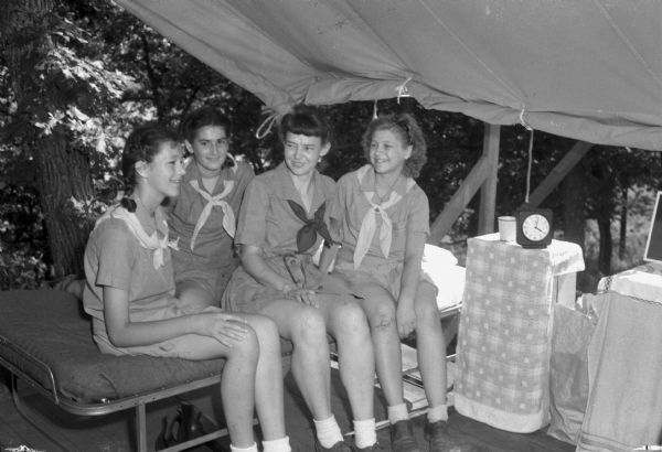 Three girl scouts and the camp director chatting while sitting on a cot in a tent at Camp Brandenburg near Springfield Corners. Pictured left to right: Mary Jane Kleinheinz, Vivian Moreland, Camp Director Mary Maxwell, and Arlene Mickelson.