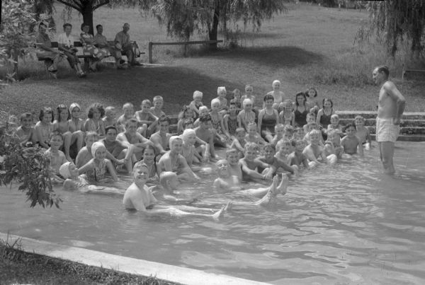 A Red Cross instructor with a children's swimming class at the Bluemounds swimming pool. The instructor is George Steiner, assistant principal at Madison West High School, and director of the Red Cross water safety and water safety program.