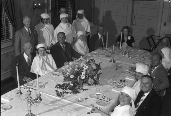 Eight couples and one woman with a corsage gathered around a dining table in the McNeel home. Eight women are wearing matching white capes and head scarves. Possibly Red Cross workers.