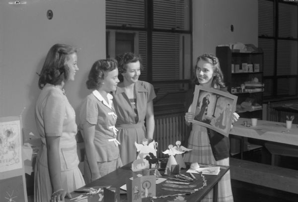 Four Junior Red Cross members with Christmas cards, tray favors, Christmas tree ornaments and recreations hall decorations made for military units and hosptials overseas.  On the extreme right is volunteer Ruth Mary Noland showing a poster to fellow volunteers, left to right:  Marianne Havey, and Virginia Bowman, and Junior Red Cross secetary, Josephine Nicholson.