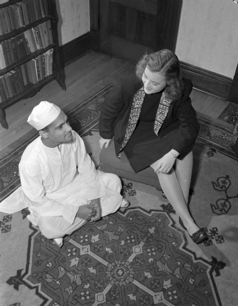 Overhead view of a woman in western attire sitting on a cushion on a floor next to a man in Middle-Eastern attire who is sitting cross-legged on the floor on an oriental carpet.