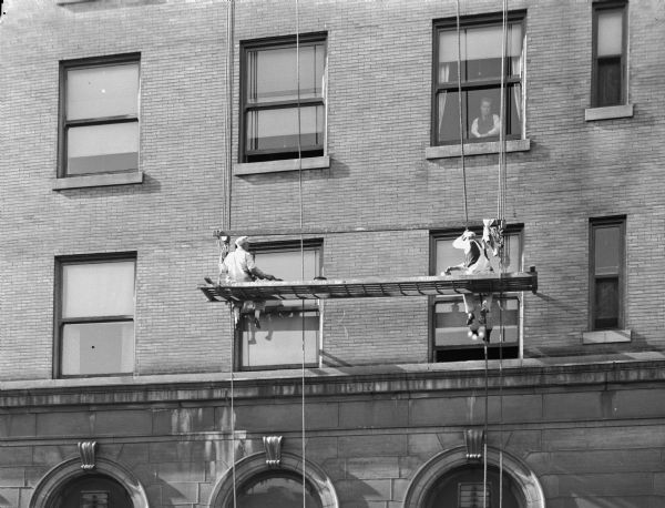 Two Capital Decorating Company employees on a scaffold painting window frames in the Park Hotel, 22 South Carroll Street. A man can be seen looking out of a window just above the painters.