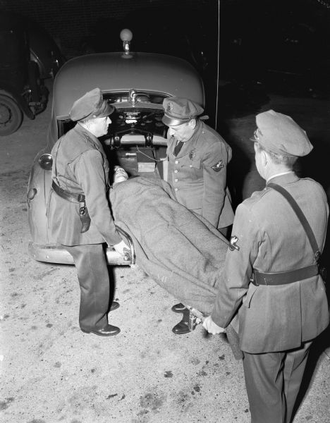 Dane County Traffic Police receiving first aid training.  Officer E.W. Kelzenberg on the left, Lieut. A.C. Pope in the middle, and Officer Ralph Conklin on the right, loading Oficer Donald Harless into the "converted" type traffic bureau ambulance.