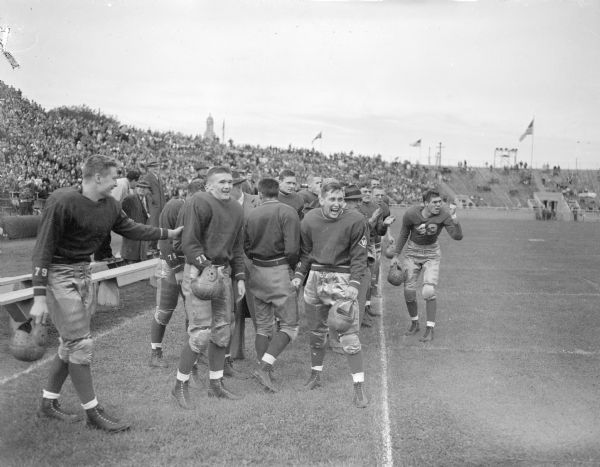 University of Wisconsin football players on the sidelines during the Wisconsin-Marquette football game.  Front row, left to right are William Koch, Jr., John English, Larry Scott, and Charles Chaney. Directly behind are Coach Harry Stuhldreher and two quarterbacks, Vern Klinzinger and Bob Engle. The Wisconsin team had just gone ahead, 20 - 6.