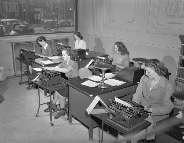 Group of five women in class at the Groves-Barnhart School for Secretaries, 502 State Street at Gilman Street. There is a large window in the background, showing pedestrians, houses and parked cars.