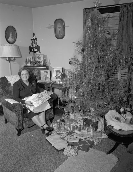 Unidentified woman seated in a chair next to a decorated Christmas tree with presents underneath. On a table next to the woman are photographs of a soldier and sailor in uniform.