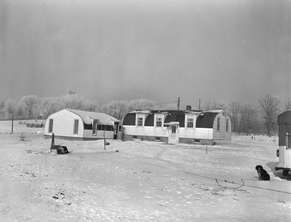 Exterior view of the Neil Young prefabricated house and garage located on Highway 12 and 18 near the Royal Airport. The building was furnished and installed by Klinke's Hatchery as part of an effort to relieve the housing shortage. A dog on a leash is sitting outside in the snow.