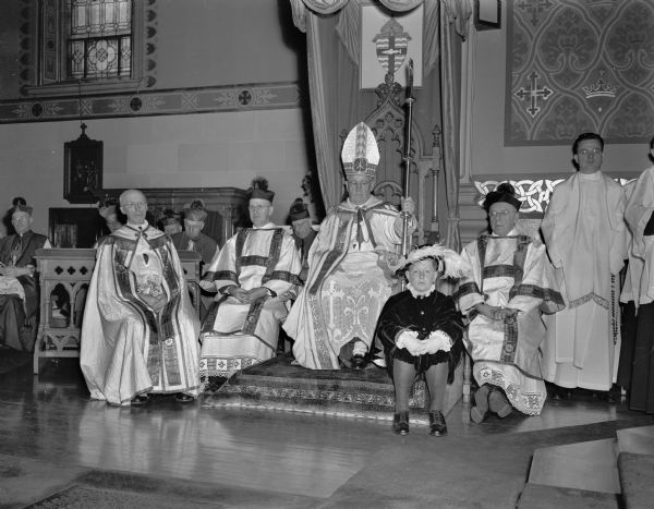 Most Rev. William P. O'Connor on his throne in St. Raphael's Cathedral during the ceremony that inaugurated him as the first bishop of Madison. L to R are: Rt. Rev. Msgr. Edward O'Reilly, Baraboo; Rt. Rev. Msgr. William Eggers of St. Bernard's, Madison (in background); Rt. Rev. Msgr. Bernard Doyle, Darlington; Bishop O'Connor on the throne, and Rt. Rev. Msgr. Joseph Delaney, Janesville, behind the page boy.