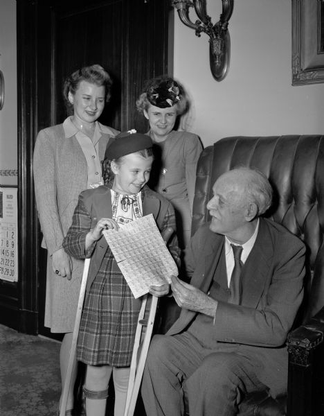 Governor Goodland being presented with a sheet of Easter Seals  by a disabled girl, Carol Schlimgen, age 9. Carol is the daughter of Mr. and Mrs. Ambrose P. Schlimgen, 4133 Meyer Avenue. Looking on are Mrs. J.L. Sonderegger, 1802 Jefferson Street, chair of the Easter Seal campaign, and Mrs. Marvin V. Bump, 3002 Oakridge Avenue, state auxiliary president of the Madison Junior Chamber of Commerce Auxiliary, sponsors of the campaign.