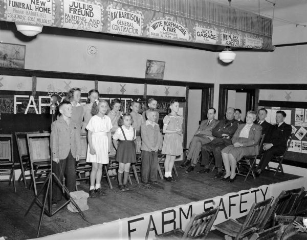 Estes School Farm Safety Program held at the Estes School on Cottage Grove Road. Ten students standing on stage with six adults sitting on stage looking on.  (Sponsored by Oscar Mayer and Company?)