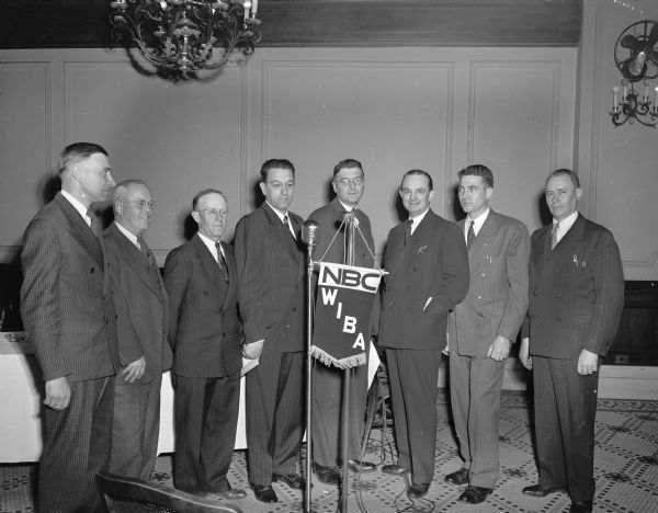 Eight men standing around a microphone in the NBC-WIBA broadcast studio including Charles A. Mittelstadt, radio commentator.