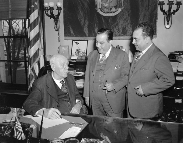 Governor Walter Goodland, seated at his desk, talking with two Mexican men.