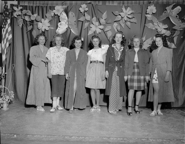 Group portrait of seven 9th and 10th grade girls modeling clothing from the Emporium Department Store for the Cottage Grove Grade School Style Show. Left to right: LaVonne Bass, Joan Hanson, Mary Ann Suchomel, Audrey Jensen, Sara Steele, Jean Hanson, and Charlotte Coffey.
