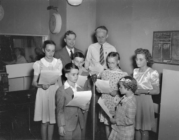Two adults and six children during an WIBA radio broadcast.  The children are standing in front of a microphone, holding a script.