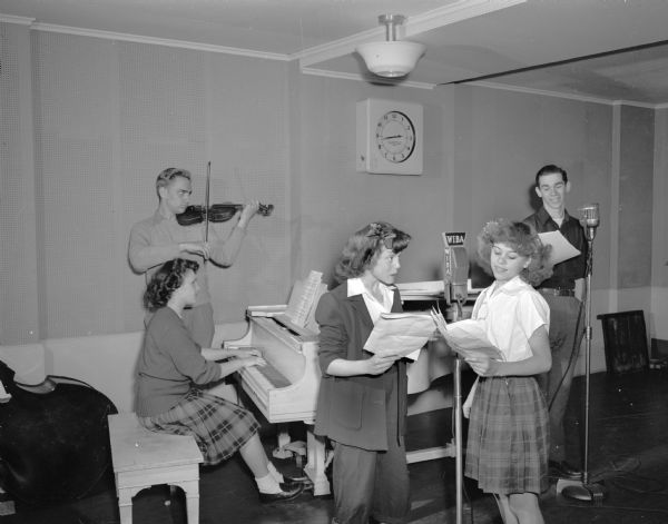 WIBA radio broadcast showing five young people; one playing the piano, one playing a violin, and three reading scripts.