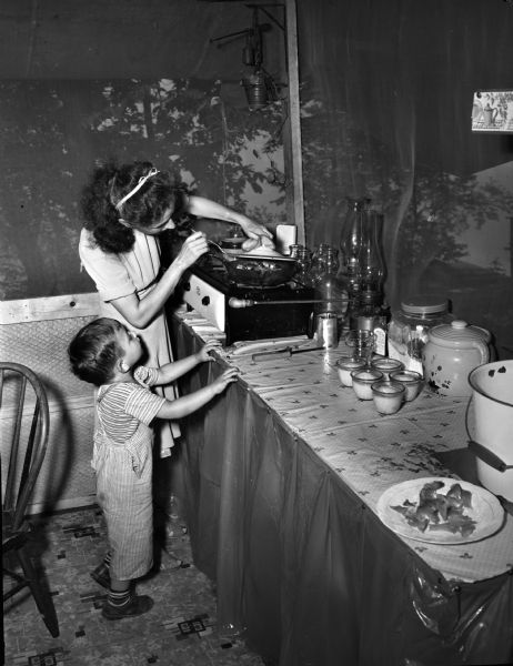 A woman and child cooking on a small gas stove in a tent at the University of Wisconsin tent colony, also known as Camp Gallistella on the south shore of Lake Mendota, west of second point.