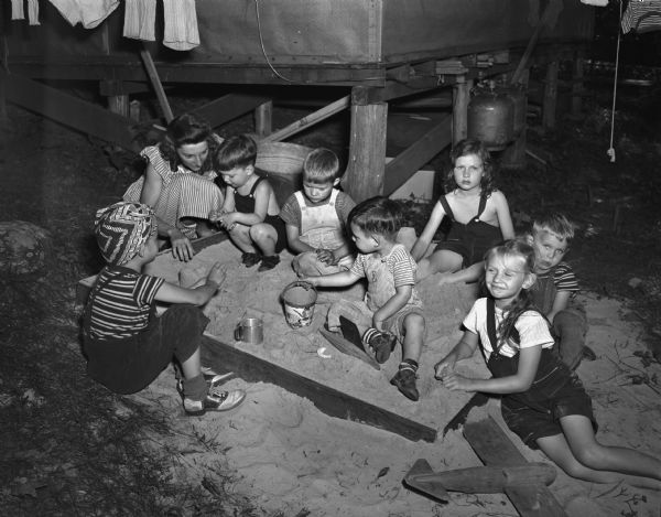 Seven children playing in the sandbox with one adult at the University of Wisconsin Tent Colony, also known as Camp Gallistella on the south shore of Lake Mendota, west of second point.