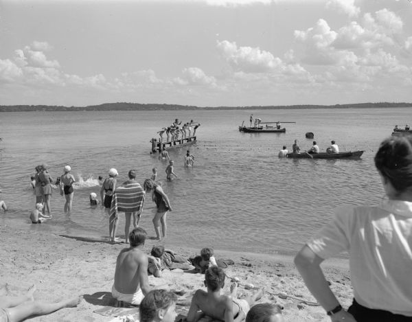 View from B.B. Clarke beach of swimmers on a raft in Lake Monona ready to start a swimming race with people in the foreground watching from the shore. Races were sponsored by Madison Board of Education Dept. of Recreation.