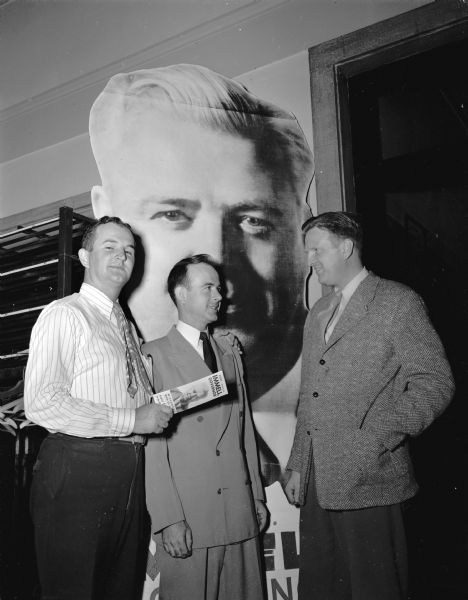 Three men, members of the University of Wisconsin Immel for Governor club. They are looking at a campaign brochure promoting Ralph Immell while standing in front of a large poster of Ralph Immell's face.